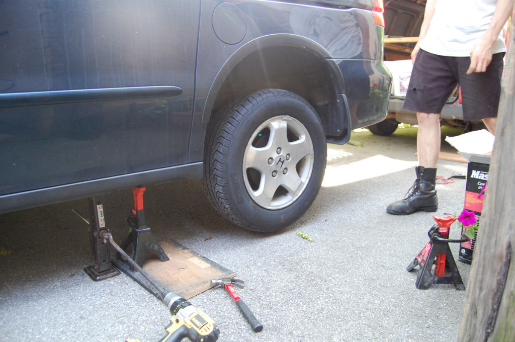 Van jacked up on the left and secured with a jack stand on the right, preparing for wheel removal.